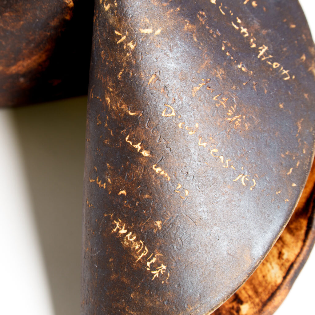 Extreme close-up of the ceramic fortune cookie surface, showing handwritten text-like scratches, dark metallic brown tones, and areas of exposed warm clay beneath the surface.