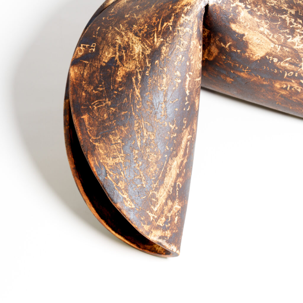 Close-up detail of a ceramic fortune cookie sculpture showing its folded edge and textured surface. Dark, smoky tones contrast with lighter raw clay marks and scratched writing across the surface.