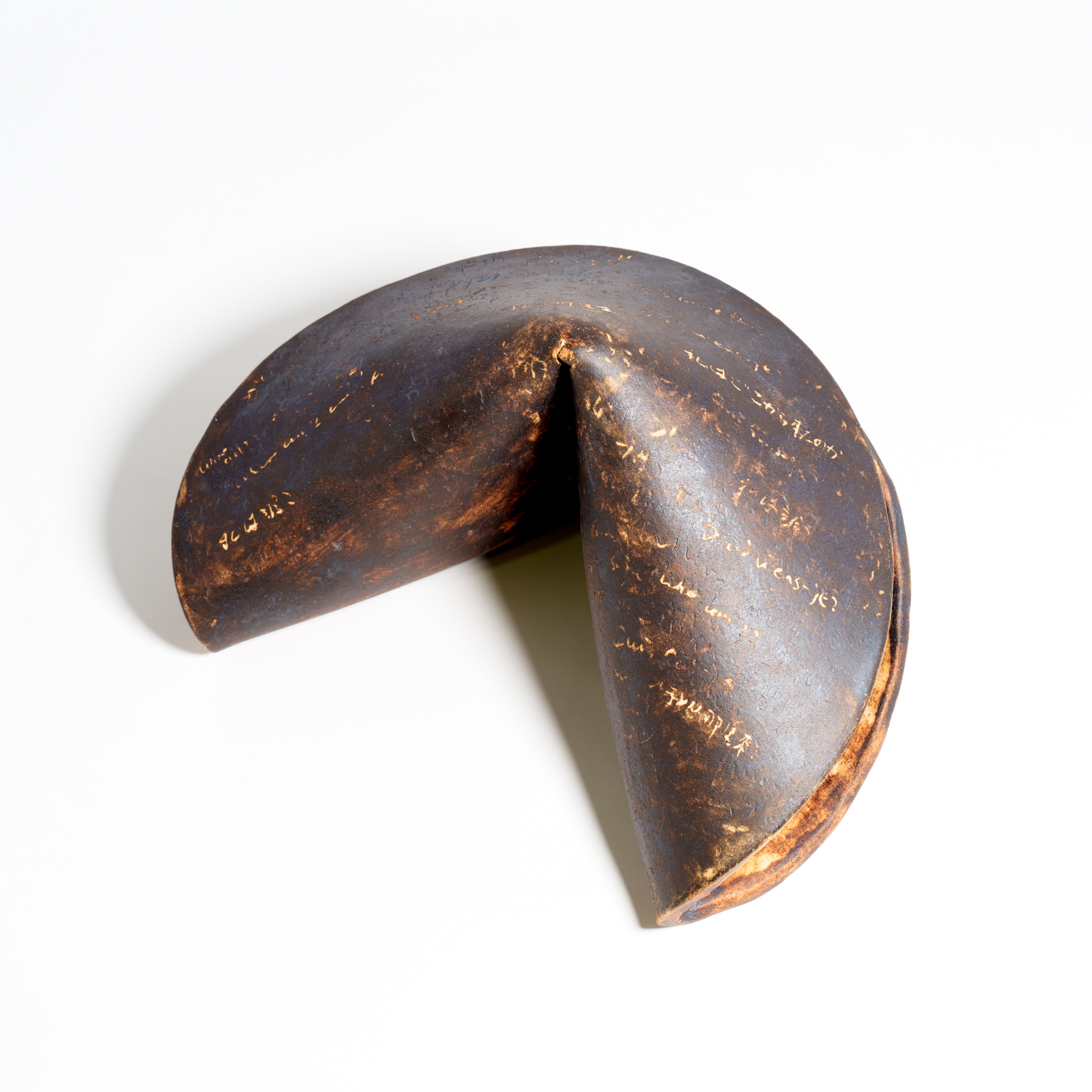 A large ceramic fortune cookie sculpture photographed from above on a white background. The surface is dark brown and black with warm, exposed clay markings and handwritten text-like scratches across the form.