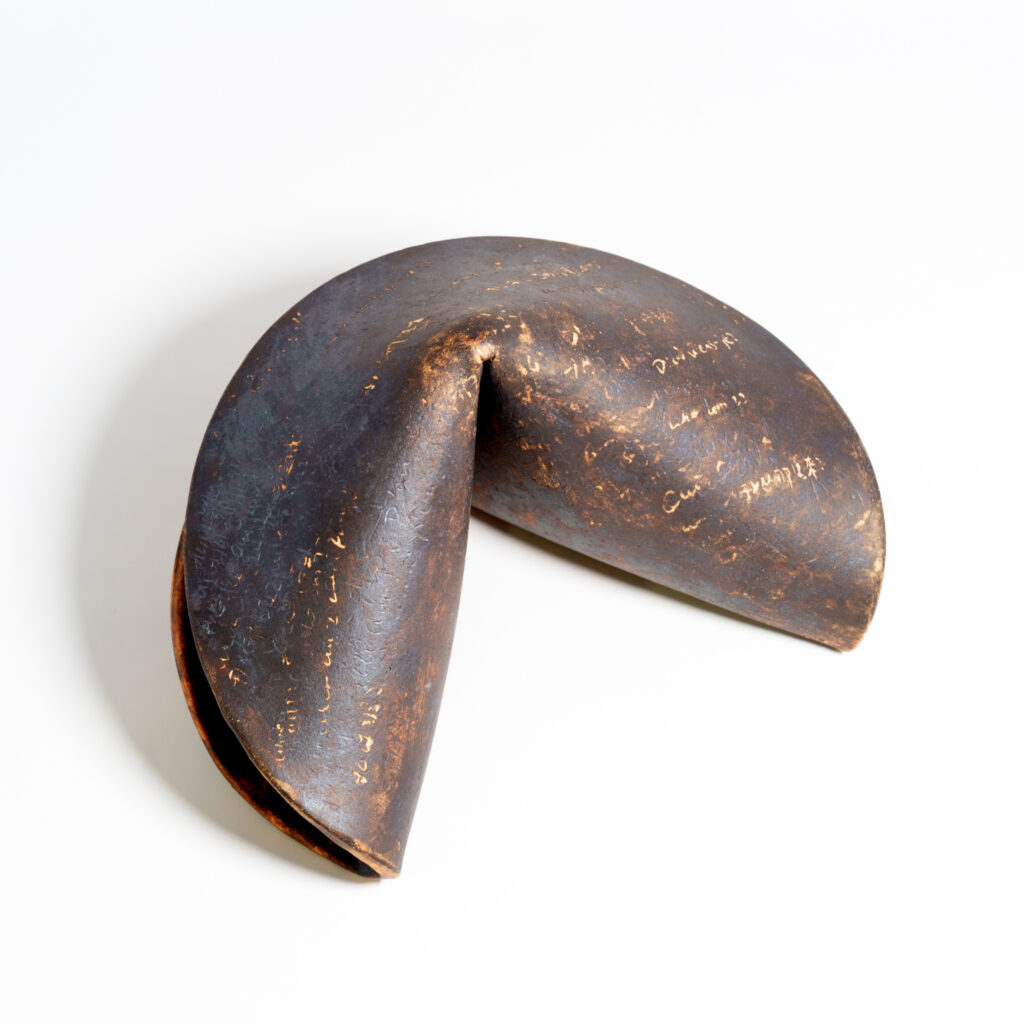 A large ceramic fortune cookie sculpture shown from a low front angle. The piece has a dark, weathered surface with warm clay highlights, scratched markings, and a hollow folded opening.