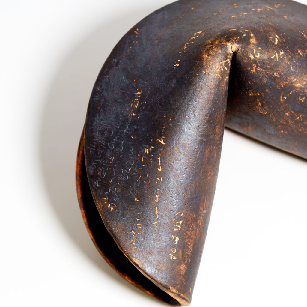 Close-up of the folded centre of a ceramic fortune cookie sculpture. The surface has a dark, burnished finish with scattered golden brown clay marks and subtle engraved text.